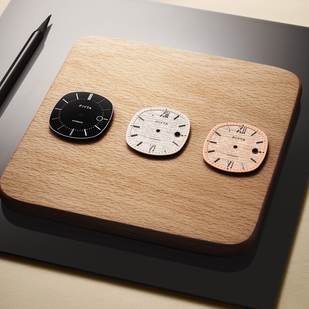 Black, light and rose-toned FIYTA Tempered dials arranged on a wooden board beside a pen.